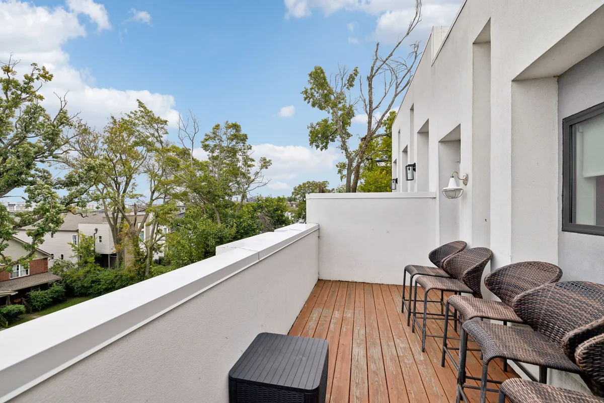 This image features a stylish balcony with a sturdy railing, woven bar stools, and a wooden deck overlooking lush green trees in the distance. The scene is bathed in natural light, suggesting either early morning or evening, and it creates an inviting space perfect for relaxing, socializing, or enjoying a drink while taking in the view.
