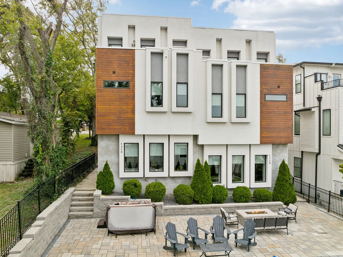 A contemporary home exterior features modern architecture with warm wood accents, a landscaped yard, and an inviting patio seating area. The patio is set for socializing, with comfortable chairs gathered around a cozy fire pit, all surrounded by lush greenery.