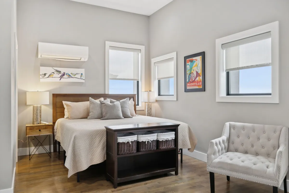 A well-appointed bedroom with a bed featuring decorative pillows, a nightstand with a modern lamp, framed artwork, and a cozy accent chair placed by the window. The soft morning light creates a calming ambiance.