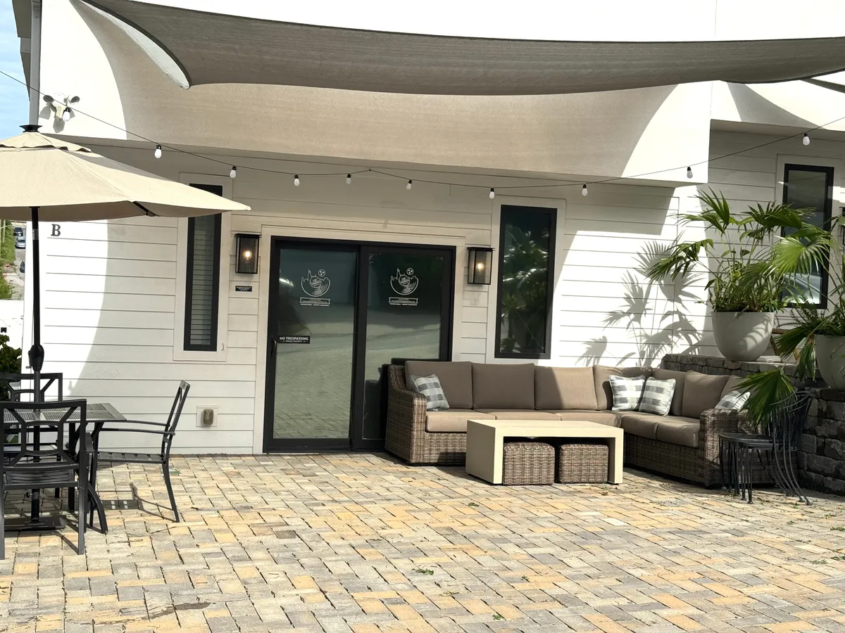 A bright exterior scene featuring a covered seating area and outdoor dining set surrounded by decorative plants. The mood is casual and welcoming, ideal for socializing, enjoying meals, or gathering for happy hour as the sun sets.