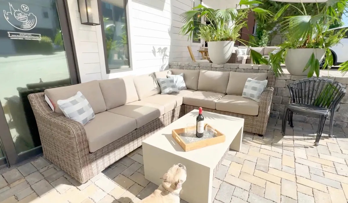 A cozy outdoor seating area with a sectional sofa, coffee table, wine bottle, decorative cushions, and potted plants. The space is designed for comfort, relaxation, and intimate gatherings, with sunlight streaming through, making it ideal for morning coffee or evening conversations.