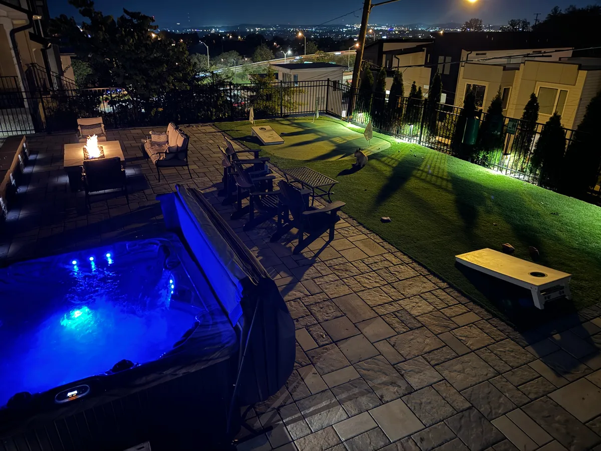 This inviting nighttime scene highlights a glowing hot tub with blue ambient lighting, a toasty fire pit with seating, and a lawn area designed for games like cornhole. The setup encourages stargazing, relaxation, and friendly competition, all under the open sky.