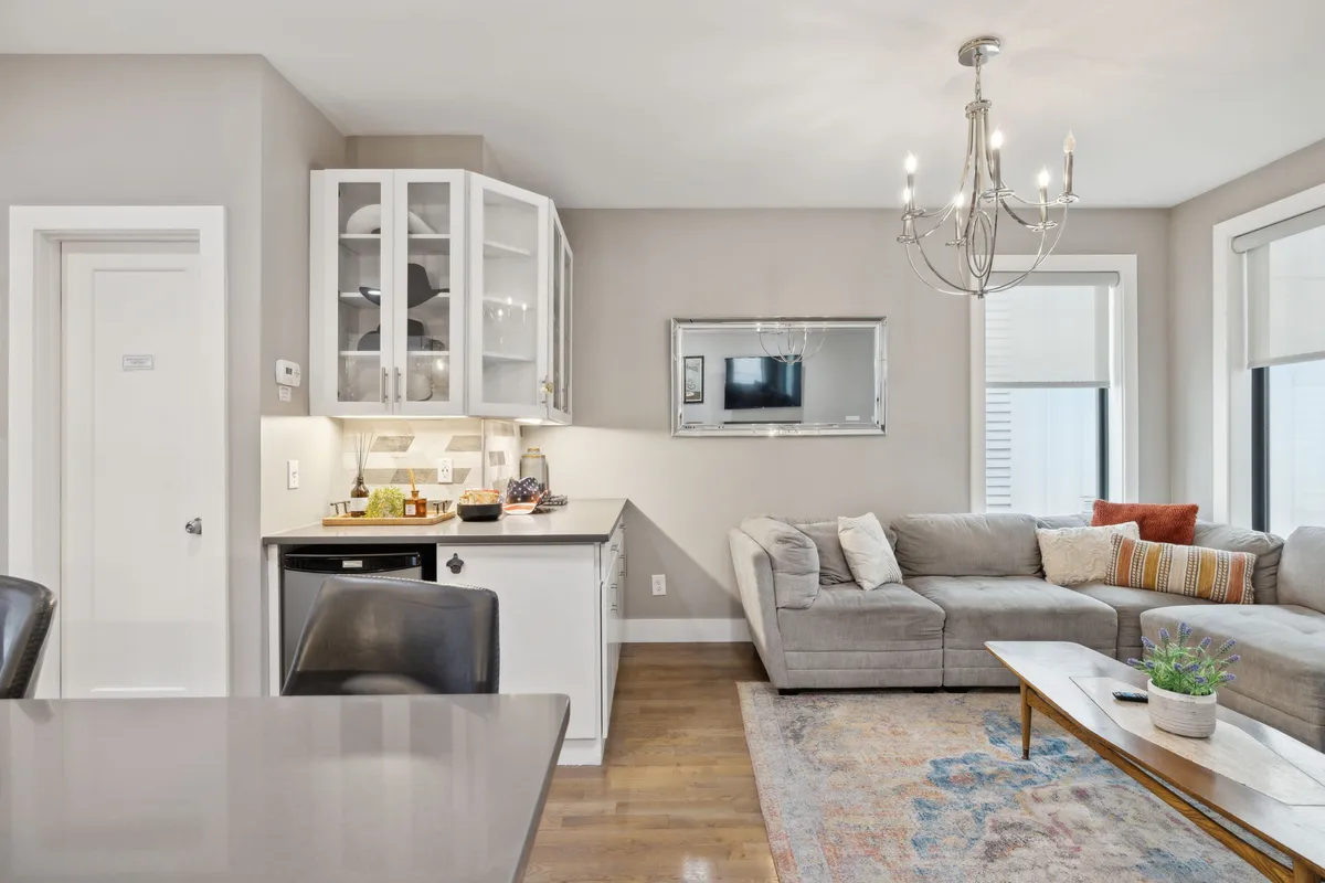 A spacious living room with a comfortable sectional sofa, a dining table set beneath a chandelier, and a view into the kitchen cabinetry. The abundance of natural light and the stylish yet inviting decor make this an ideal space for both group relaxation and shared meals.