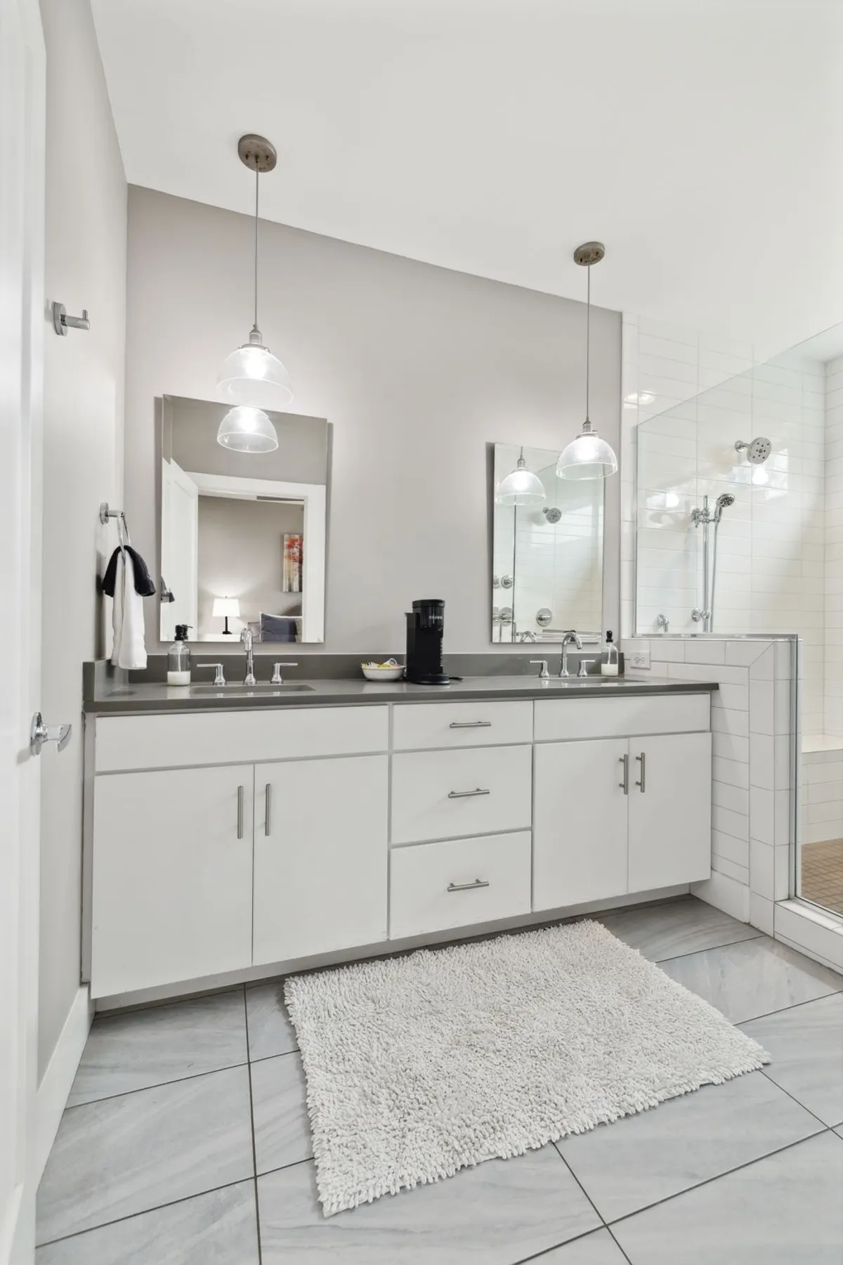 A tranquil, modern bathroom featuring a double sink with sleek fixtures, expansive mirrors, a shower area with glass doors, pendant lights, and a plush rug. The space feels inviting and perfect for self-care rituals.
