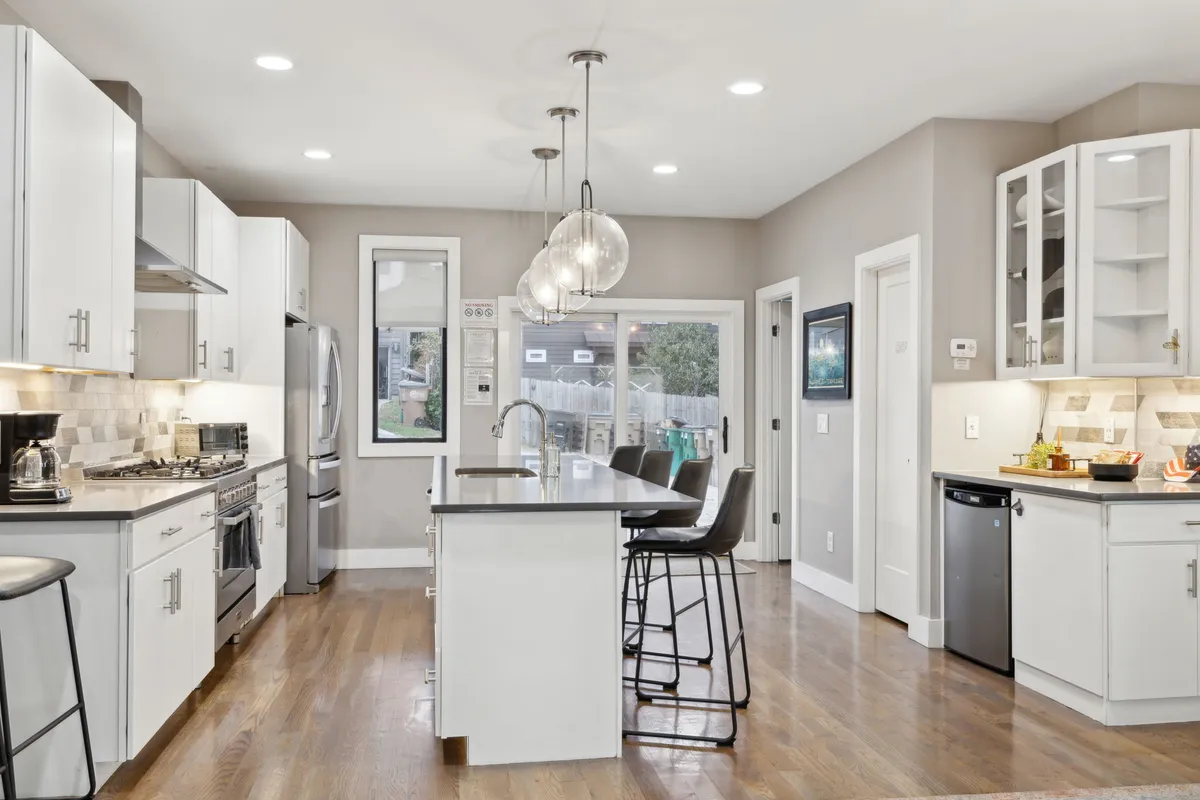 A contemporary kitchen with an island featuring seating, modern appliances, bright pendant lighting, and windows that offer a glimpse outdoors. The island setup encourages conversation and casual dining, while the light-filled space feels both inviting and functional.
