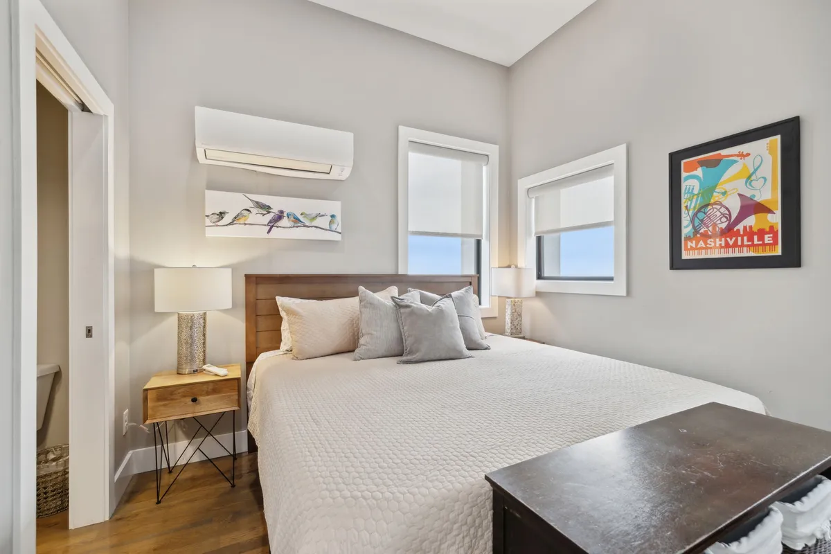 A spacious bedroom featuring a plush king-sized bed dressed in crisp linens, with soft natural light pouring in from large windows. The room includes stylish artwork above the headboard, a sleek nightstand with a contemporary lamp, and serene decor that evokes relaxation.